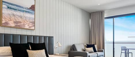 A modern hotel room with a large bed, a sofa, a dining area, and a balcony with a view.