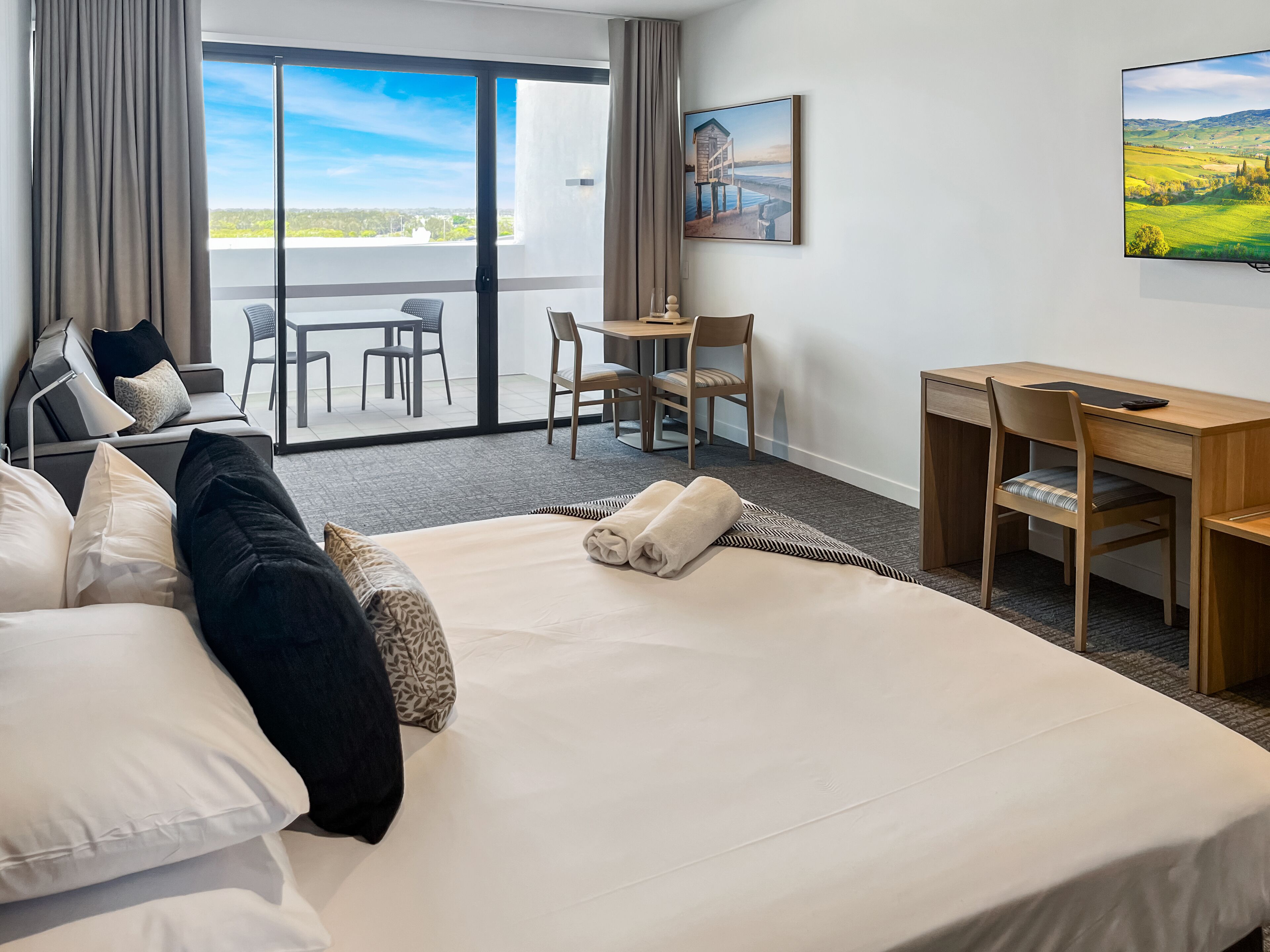 Quarto de hotel com uma cama grande, uma escrivaninha, uma cadeira, uma mesinha e vista para o exterior.