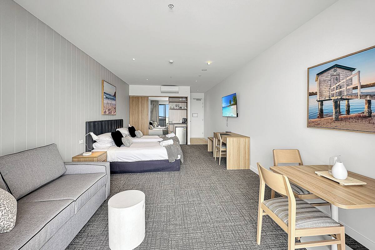 Habitación de hotel con cama, sofá, mesa de comedor con sillas, televisor y un cuadro en la pared.