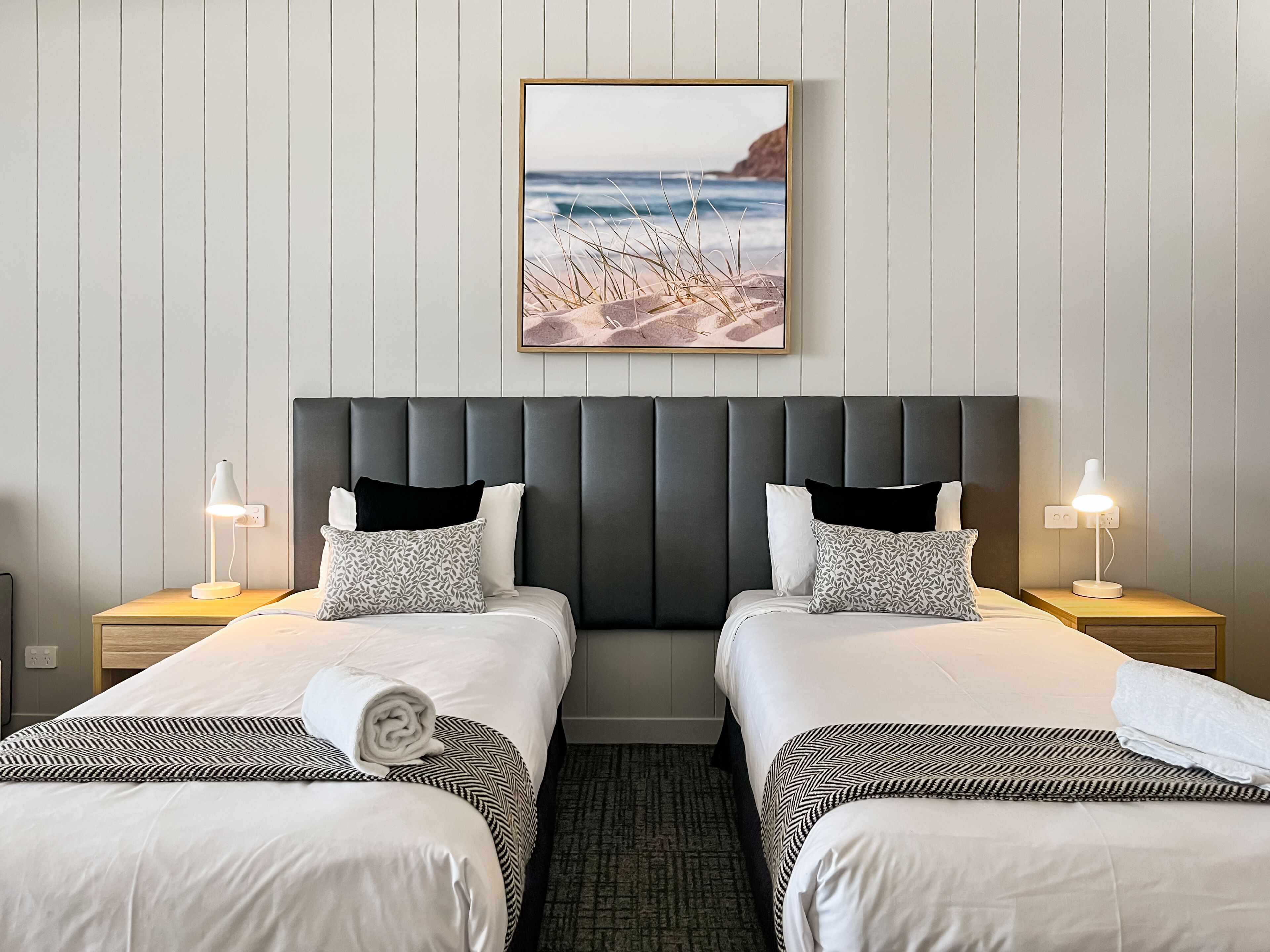 Una habitación de hotel con una cama grande, dos mesitas de noche, un cabecero y un cuadro con una playa.