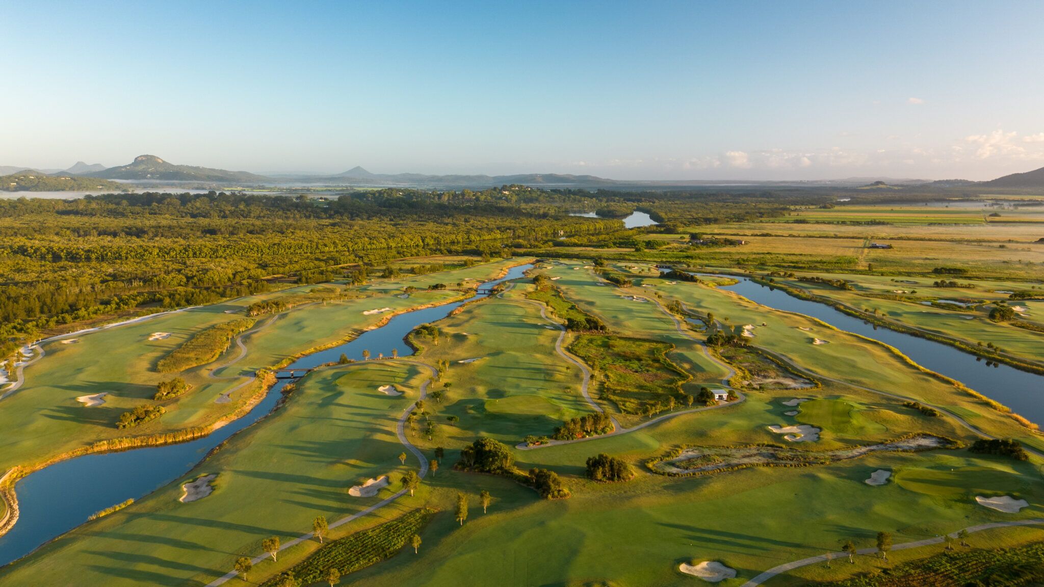 Ein Golfplatz mit mehreren Wasserhindernissen, Sandbunkern und einem Clubhaus in der Ferne.