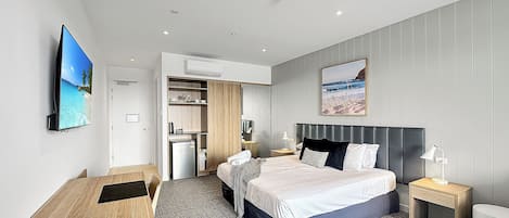 A modern hotel room with a bed, bedside table, and a desk with a chair. There is a wardrobe, a small kitchenette, and a TV mounted on the wall.