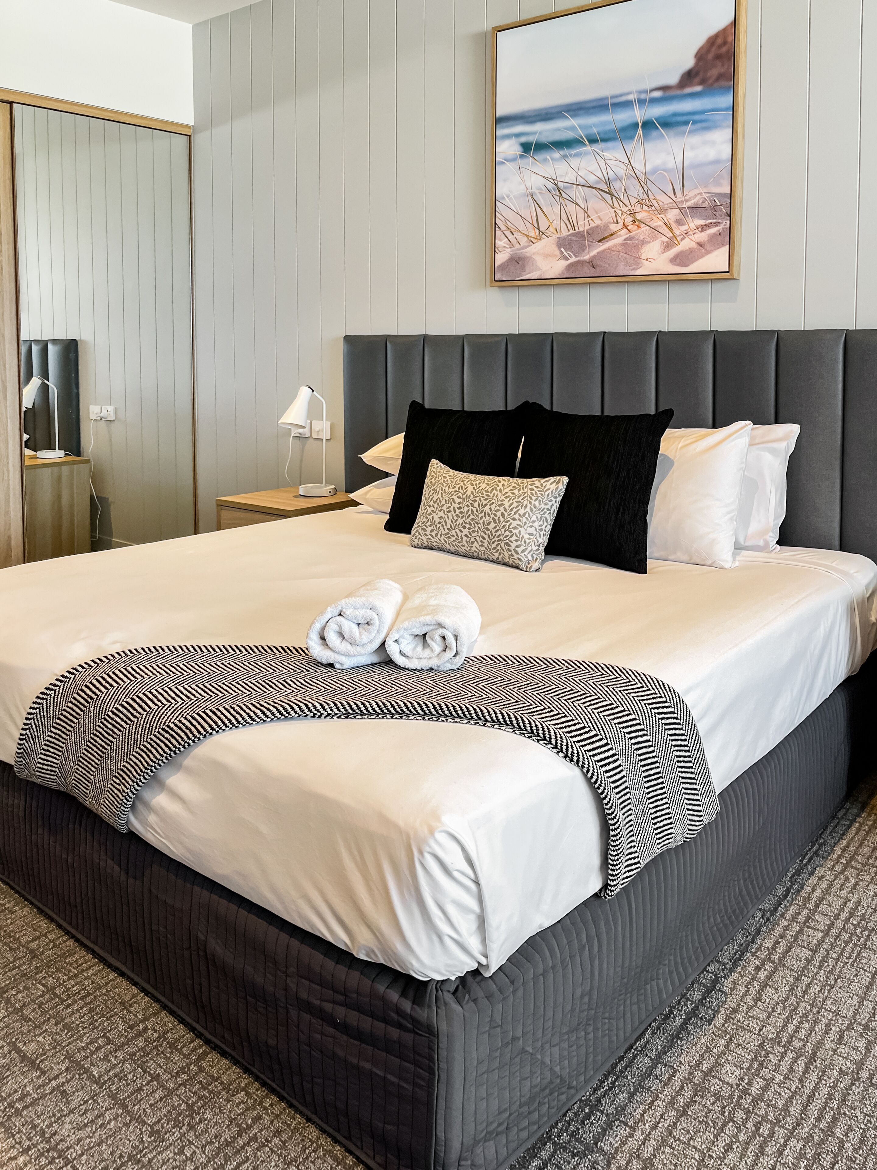 A neatly made bed with white linens, a black and white patterned throw, and rolled towels.