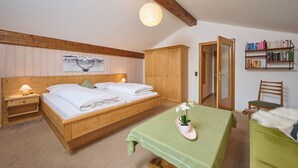 Un dormitorio de madera con una cama, mesitas de noche, una mesa verde con una planta, una estantería y una puerta que da a otra habitación.