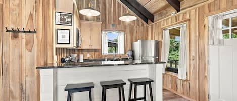 A wooden interior with a kitchen area featuring a bar stool seating, a refrigerator, and a window with curtains.