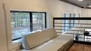 Ein Zimmer mit einer fest eingebauten Couch, großen Fenstern mit Blick auf einen Wald, Holzböden und einer Decke mit sichtbaren Balken.