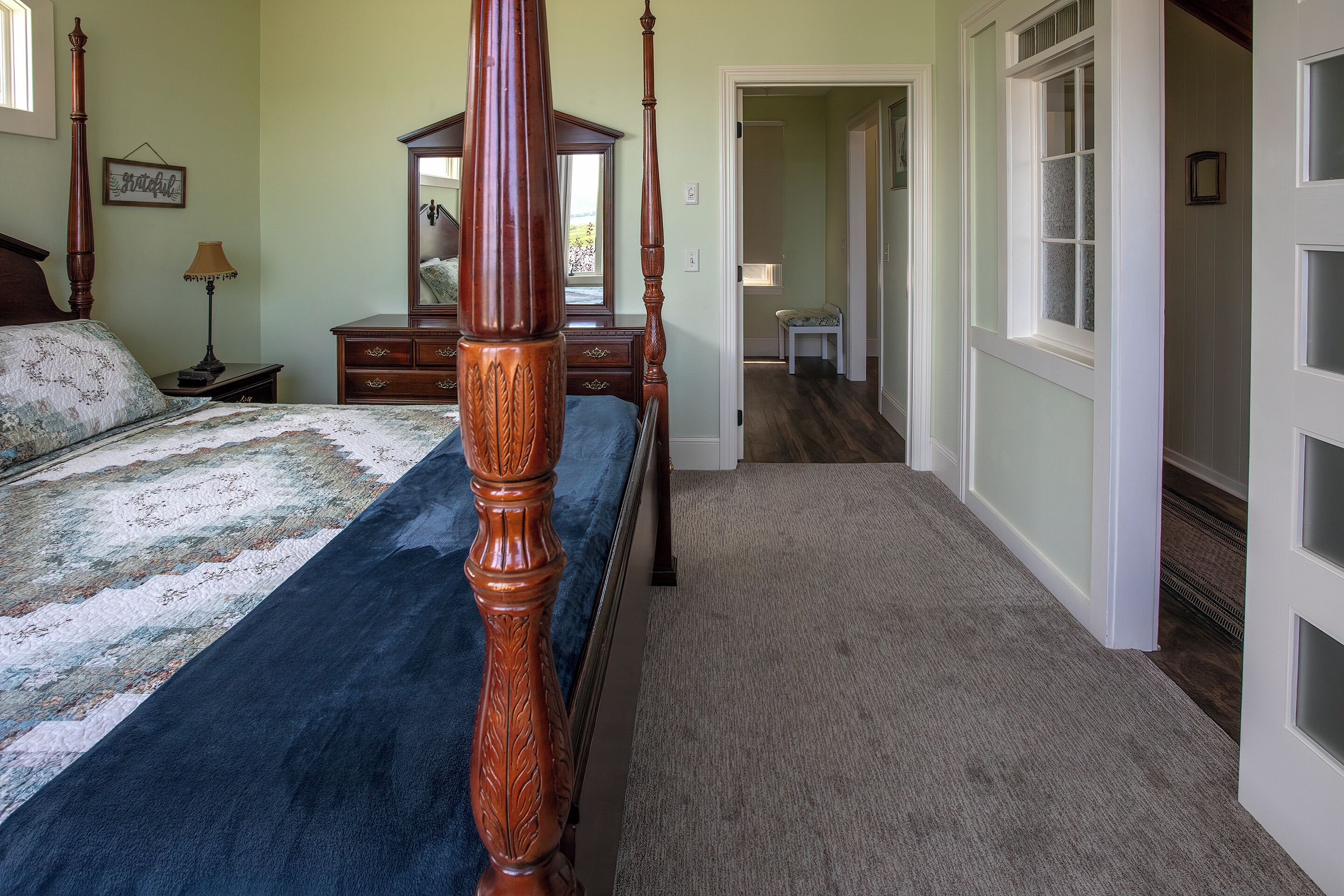 A bedroom with a four-poster bed, a nightstand, a dresser, and a mirror. There is a door leading to another room.