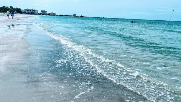 Une plage avec une eau claire, un rivage sablonneux et des bâtiments en arrière-plan.