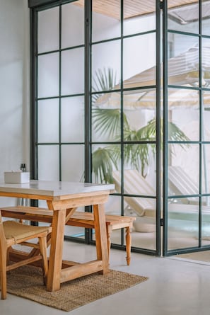 Um quarto com uma mesa e cadeiras de madeira, um tapete tecido e uma janela grande com vista para palmeiras.