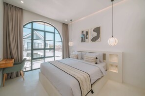A modern bedroom with a large window, a bed with white bedding, a desk, and two wall-mounted light fixtures.