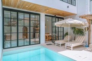 A modern indoor pool area with clear glass walls, wooden ceiling, and lounge chairs with striped cushions.