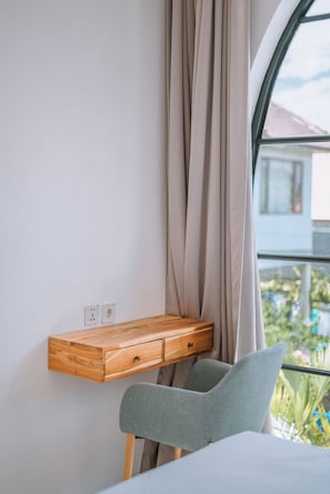 Ein Schlafzimmer mit einem Nachttisch aus Holz, einem grauen Sessel, einem Fenster mit Vorhängen und Blick auf Grünflächen.