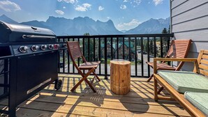 Een balkon met een barbecue, houten stoelen, een klein houten tafeltje en uitzicht op bergen.