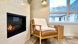 Een moderne woonkamer met een open haard, een leren fauteuil en een groot raam met uitzicht op gebouwen.