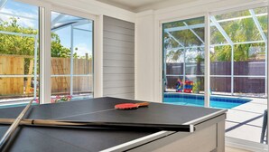 Ein Tischtennistisch in einem Raum mit großen Fenstern, die auf einen Garten mit Pool blicken.