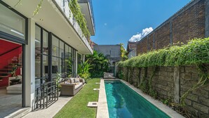 A modern outdoor pool area with a glass-enclosed patio, a small seating area, and a brick wall with climbing plants. - Villa Jungalow 2BR Pool Villa Near Beach Café del Mar & Padel (Canggu)