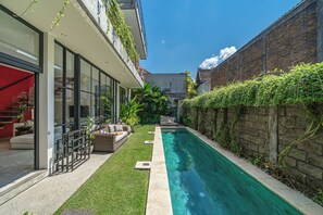 A modern outdoor pool area with a glass-enclosed patio, a small seating area, and a brick wall with climbing plants. - Villa Jungalow 2BR Pool Villa Near Beach Café del Mar & Padel (Canggu)