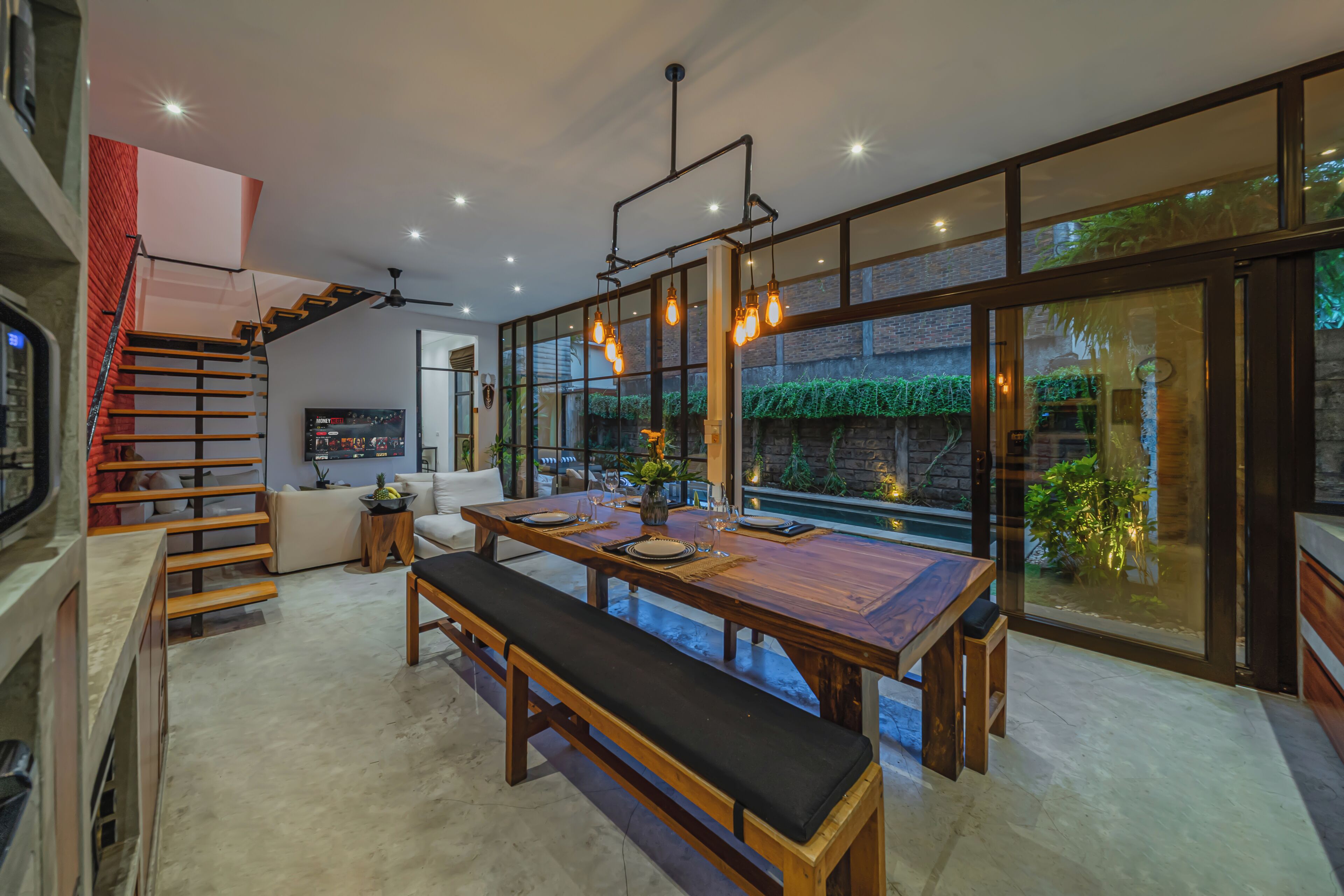 A modern dining area with a wooden table, bench, and hanging light fixtures.