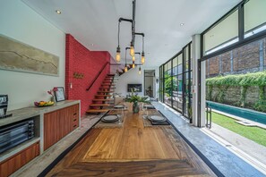 A modern kitchen with a wooden island, a red brick wall, and a dining area with a long table set for four. - Villa Jungalow 2BR Pool Villa Near Beach Café del Mar & Padel (Canggu)