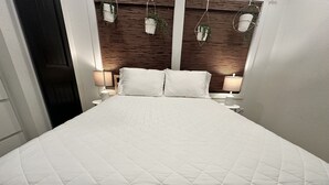 Una cama bien hecha con un edredón blanco acolchado, dos lámparas de noche y un cabecero de madera con macetas colgantes.