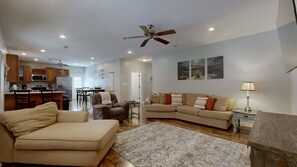 A spacious living area with a large sofa, a smaller sofa, and a dining table. There is a kitchen area with wooden cabinets and a ceiling fan.