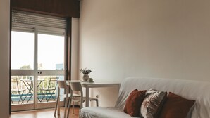 Un salon moderne avec un canapé blanc, une table à manger avec des chaises, un luminaire suspendu et une vue dégagée sur l’extérieur grâce à une grande fenêtre.