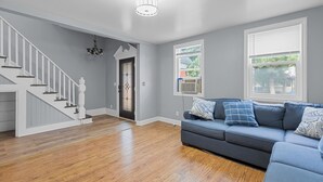 A spacious living room with hardwood flooring, a gray sofa with patterned cushions, a staircase with white railings, and a chandelier.