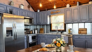 A kitchen with grey cabinets, stainless steel appliances, a wooden dining table with breakfast items, and a window with blinds.