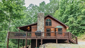 A wooden cabin with a stone chimney, a deck, and a view of the surrounding forest.