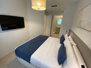 Une chambre à coucher avec un lit, un téléviseur fixé au mur et une salle de bain visible à travers une porte ouverte.