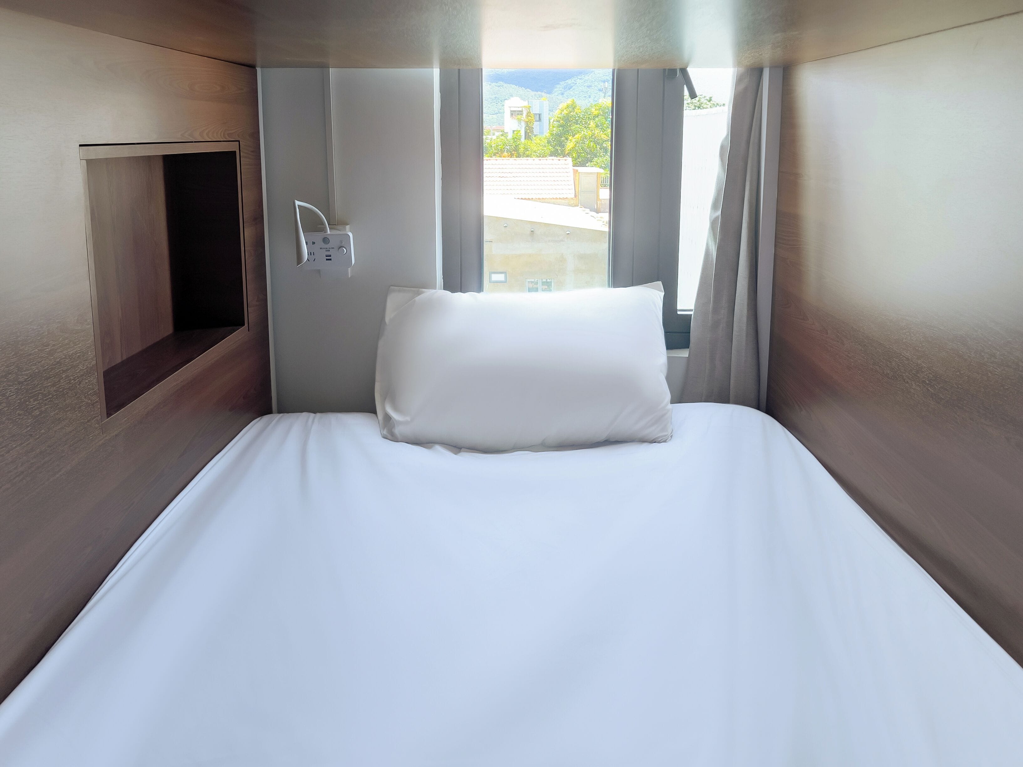 Una habitación de hotel con cama, ventana con cortinas y panel de control en la pared.