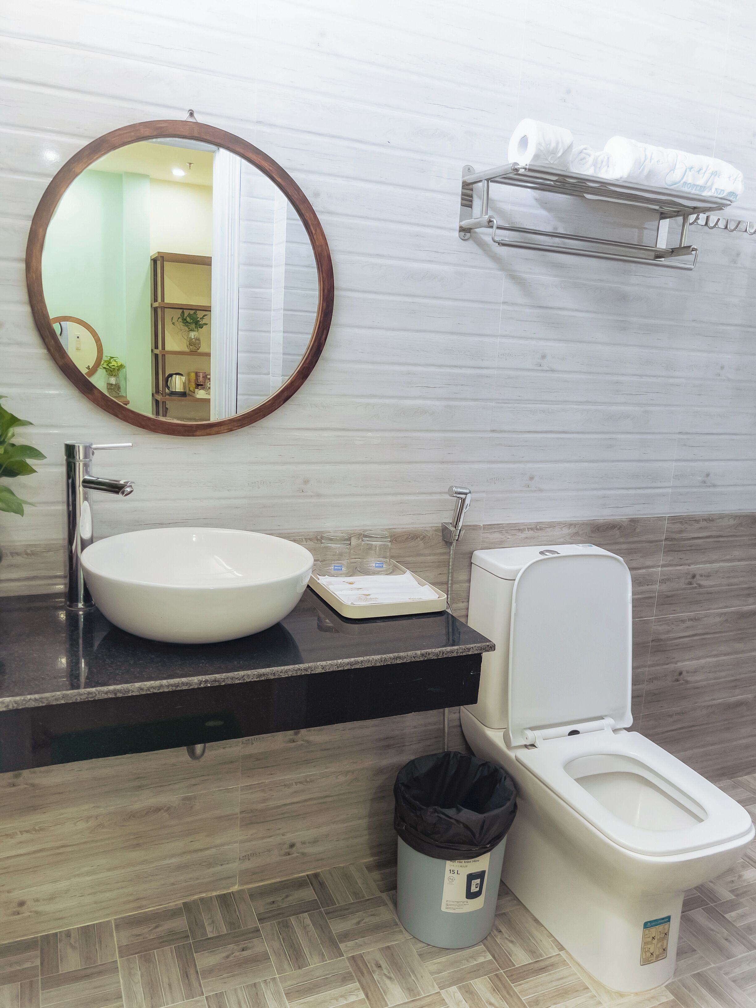 A bathroom with a round mirror, a white sink, a toilet, and a trash bin.