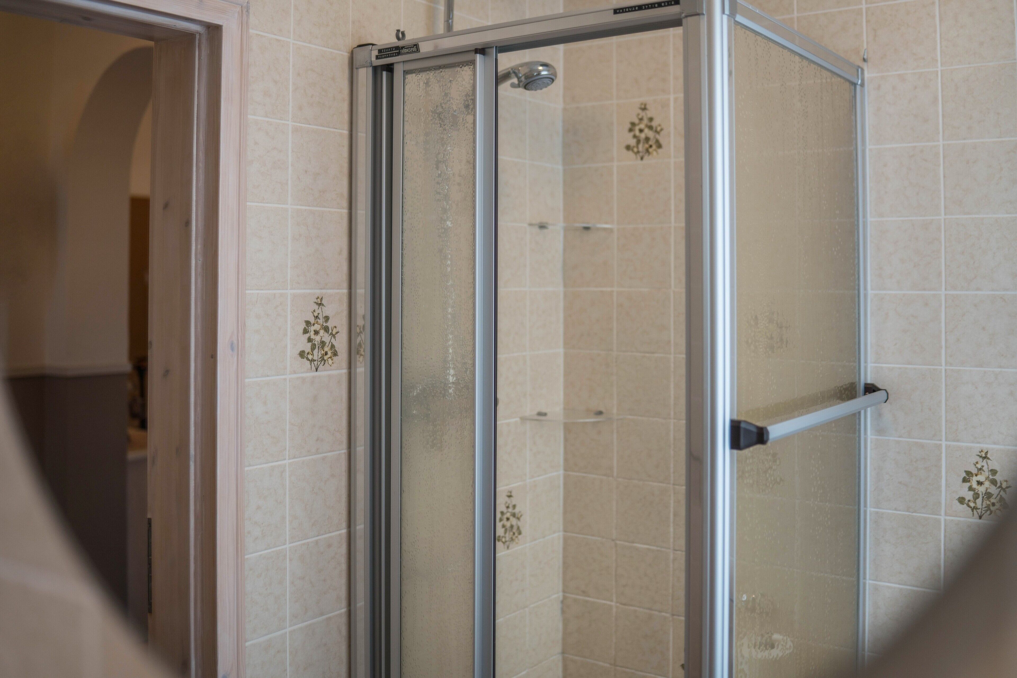Une douche fermée par des parois de verre, avec une structure métallique, des murs recouverts de carreaux beiges et une pomme de douche visible.