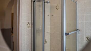 Une douche fermée par des parois de verre, avec une structure métallique, des murs recouverts de carreaux beiges et une pomme de douche visible.