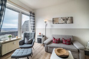 A modern living room with a grey sofa, a grey armchair, a wooden side table, a radiator, a floor lamp, and a window with a view.