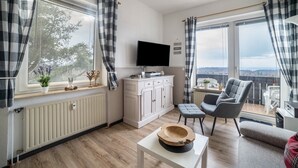 A modern living room with a flat-screen TV, a grey armchair, a sofa, a small table with a hat, and a view of the city through large windows. - Apartment 'Harzperle Nr 6' with Mountain View, Balcony and Wi-Fi (Braunlage)