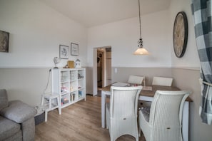 Un coin repas avec une table en bois, des chaises en osier blanches, une étagère blanche contenant divers objets et un luminaire suspendu.