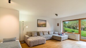 Un salon moderne avec un canapé d’angle, une lampe sur pied et une grande fenêtre donnant sur un paysage verdoyant.