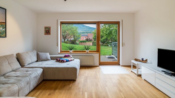 Un salon moderne avec un canapé, un téléviseur et une grande fenêtre donnant sur un jardin et des immeubles.