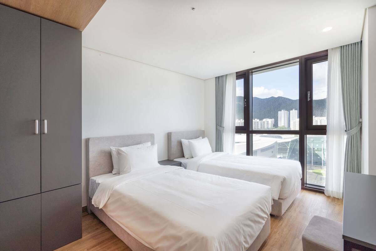 Una habitación de hotel moderna con dos camas, un ventanal grande con vista a la ciudad y un armario gris.