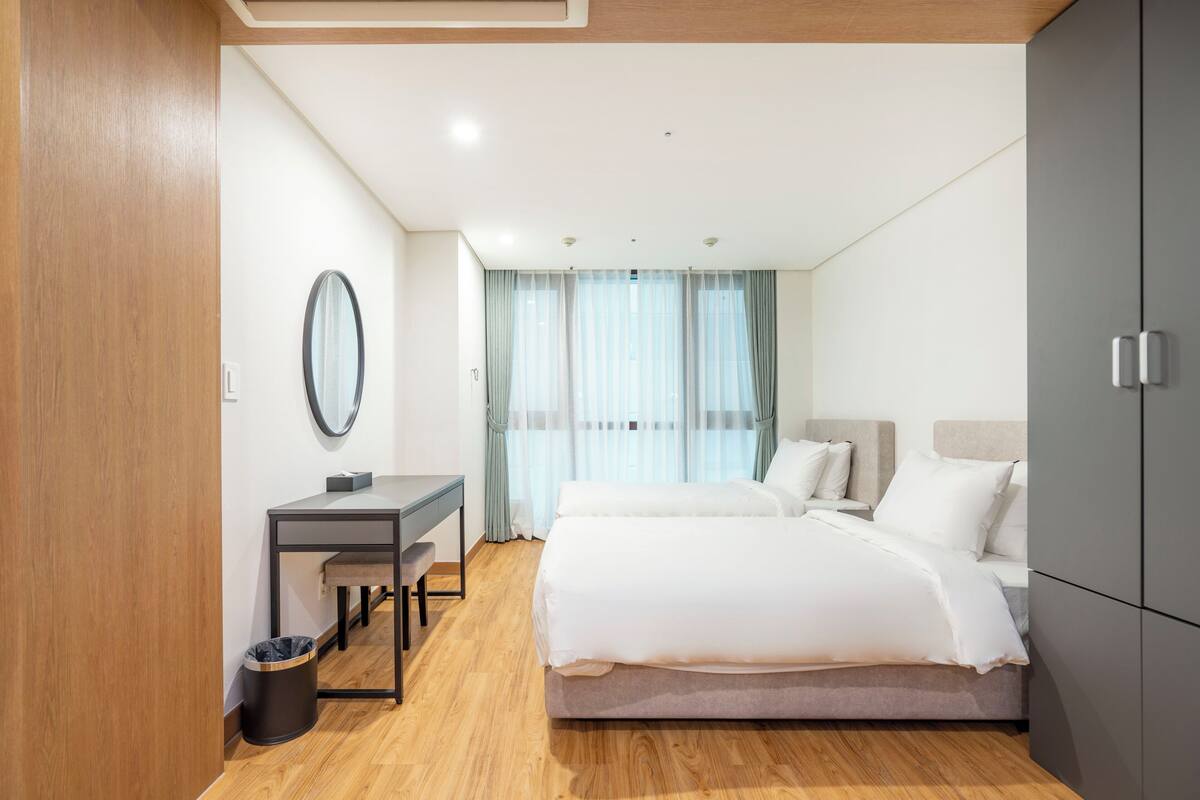 Una habitación de hotel moderna con piso de madera, un escritorio con silla, una cama con ropa blanca y un armario.