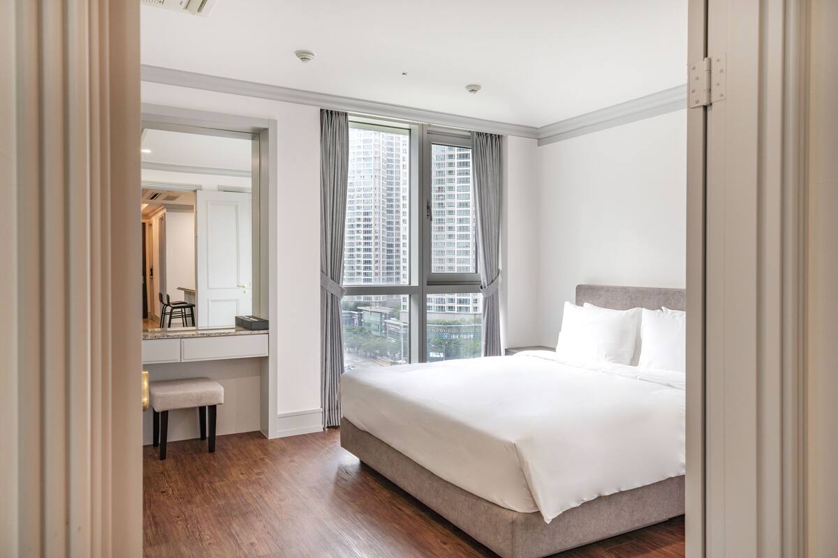 Un dormitorio moderno con una cama grande, un escritorio con silla y vistas a un paisaje urbano a través de una ventana.