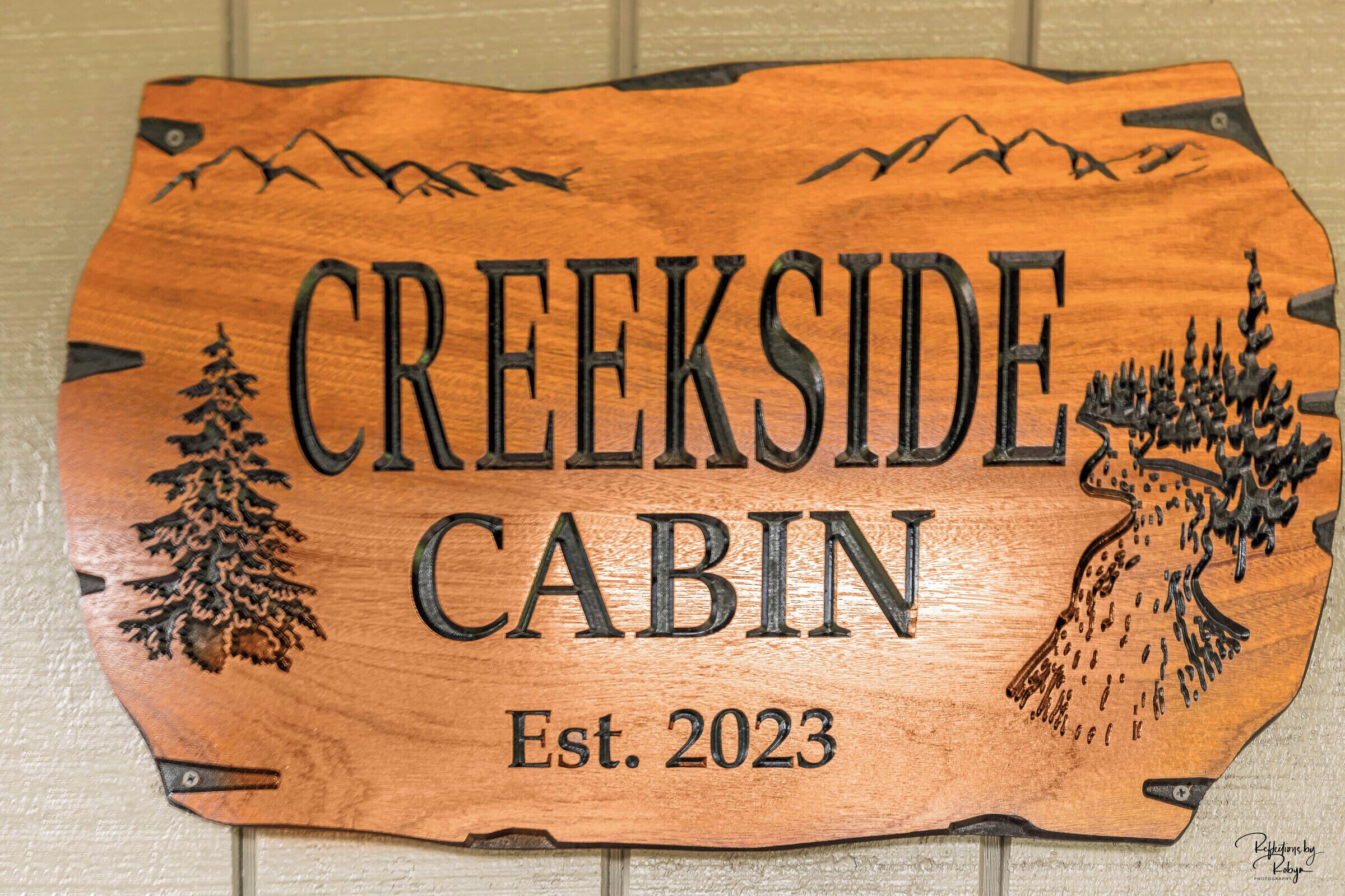 A wooden sign with the text "CREEKSIDE CABIN" and "Est. 2023" indicating the name and establishment year of a cabin.