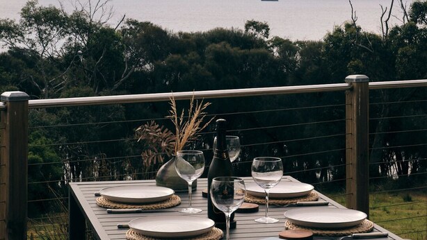 An outdoor dining table set with plates, glasses, and a bottle, overlooking a body of water and distant mountains. - Sun-drenched with spectacular views. Central location for exploring Bruny. (Alonnah)