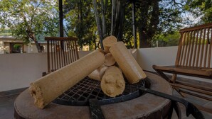 Un barbecue avec des bûches et un ustensile à long manche, entouré de chaises en bois et d’un patio couvert, avec des arbres en arrière-plan.