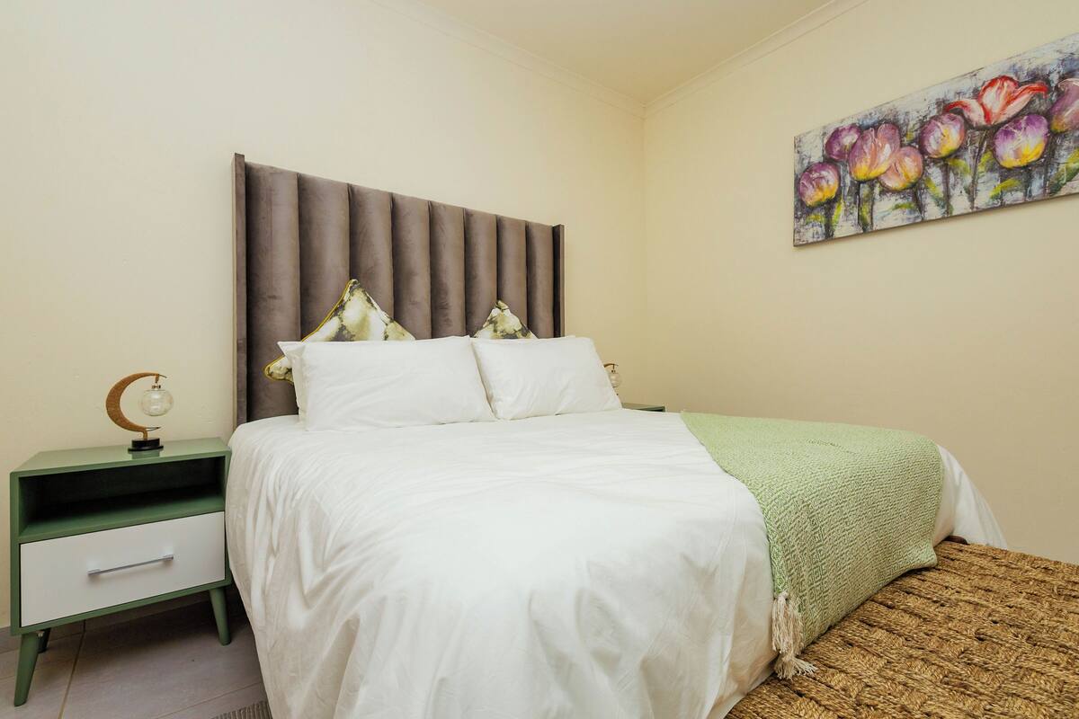 Un dormitorio con cama, mesita de noche, lámpara y un cuadro en la pared.