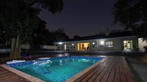 Une piscine rectangulaire avec une terrasse en bois, entourée d’arbres, et un bâtiment dont les fenêtres sont illuminées en arrière-plan.