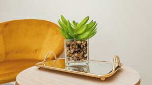 Une table ronde en bois, sur laquelle se trouve un vase en verre contenant des plantes vertes et des petits cailloux, un plateau doré et, au fond, une chaise jaune.