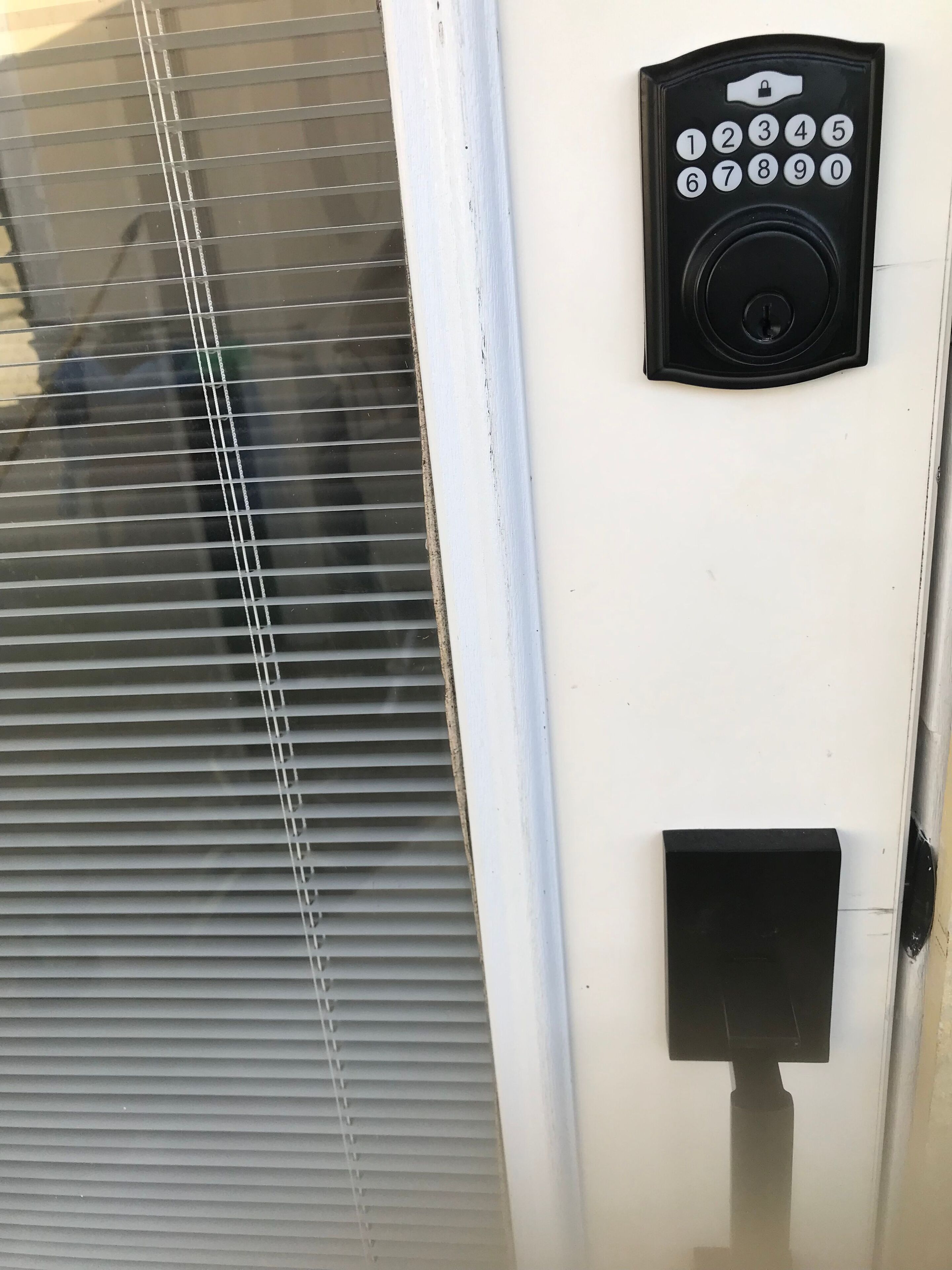 A door with a keypad lock, a window with blinds, and a small black panel below the lock.