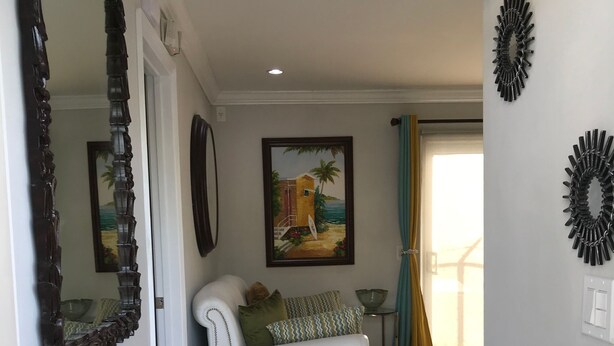 A well-lit hallway with a white sofa, a painting of a beach scene, and a mirror on the left wall. - The Haven’s hideaway (Freeport)